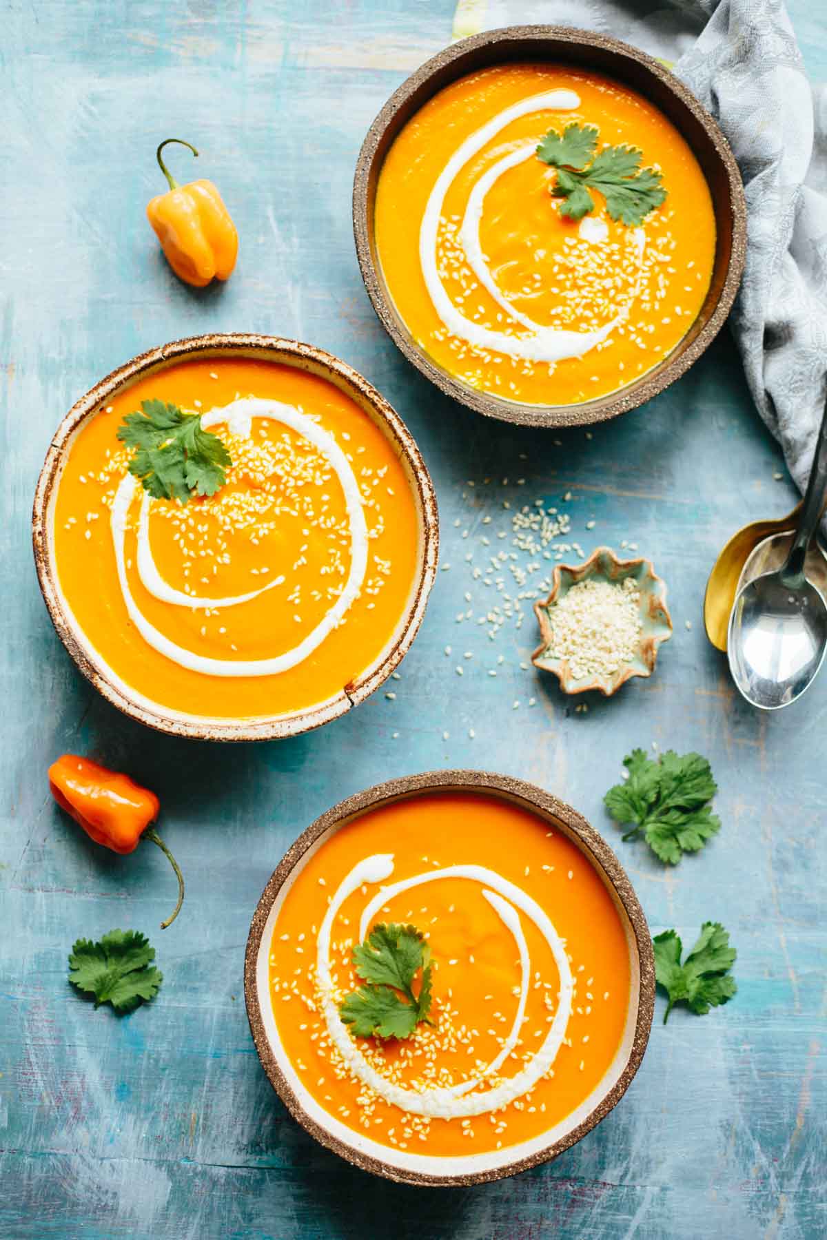 Three bowls of spicy carrot soup topped with yogurt swirls, sesame seeds, and cilantro leaves on a blue tabletop with habanero peppers nearby.
