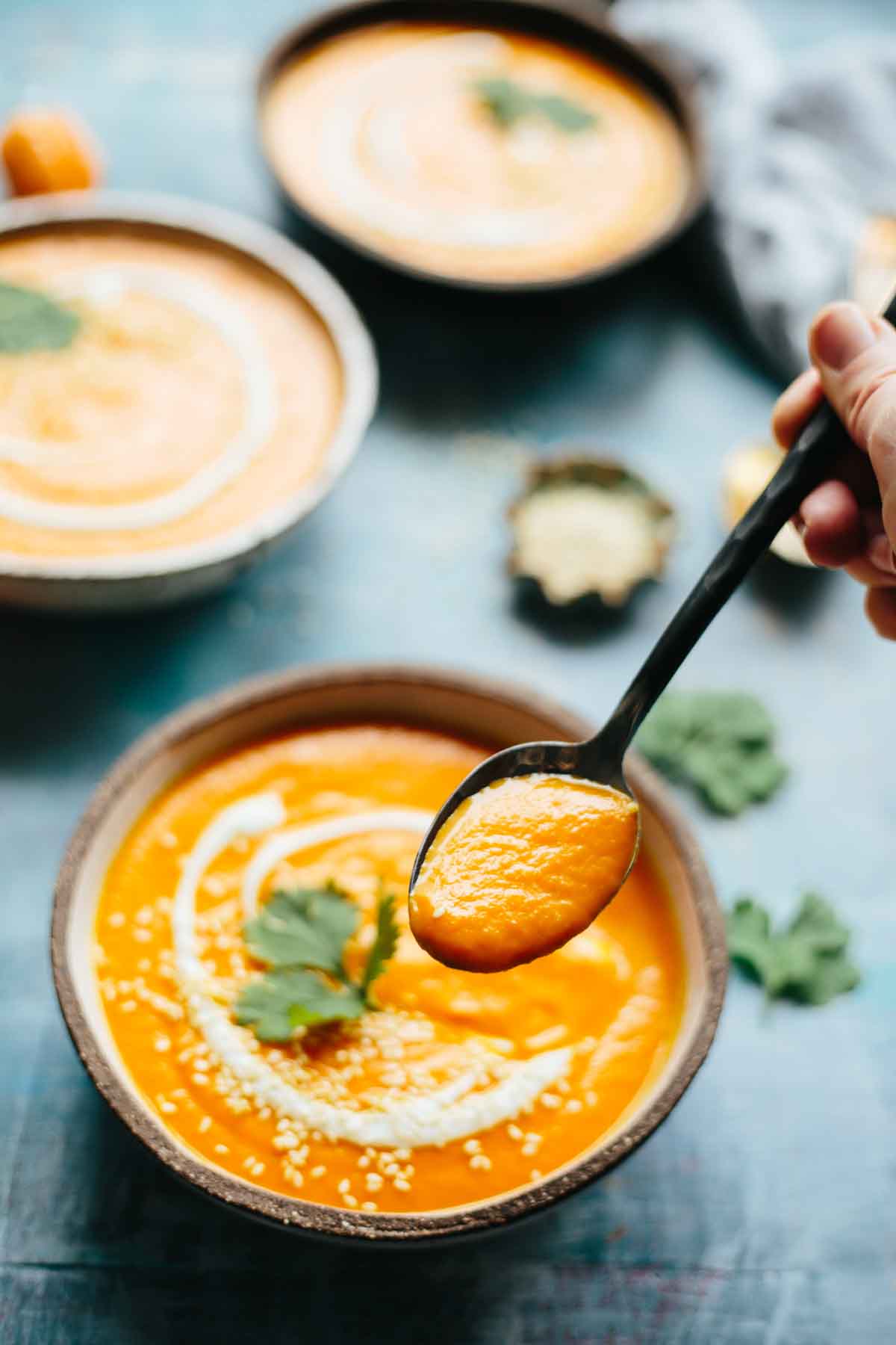 Hand holding a spoonful of spicy carrot soup over a rustic bowl topped with yogurt and cilantro.