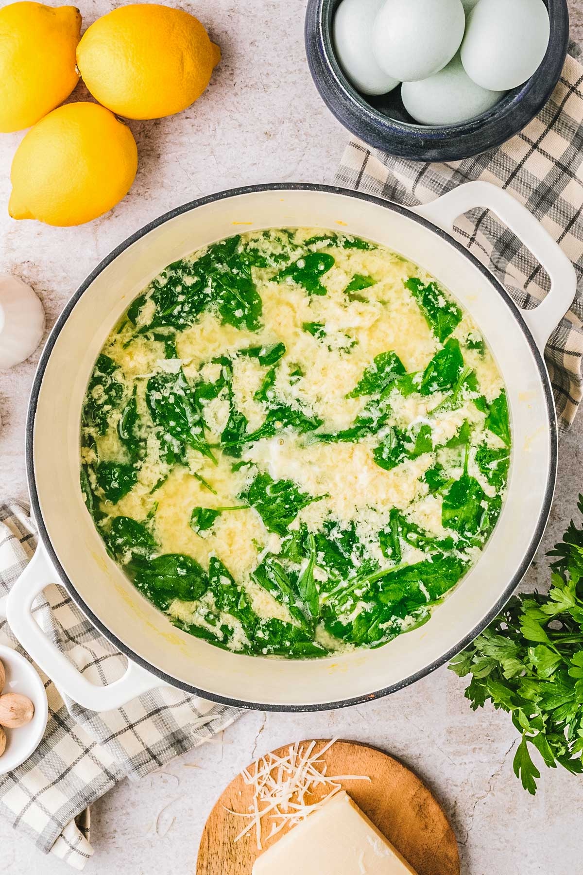 Large pot of stracciatella soup with cooked egg strands and spinach leaves, with fresh lemons and eggs in the background.