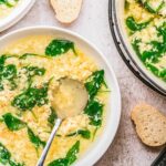 Close-up of stracciatella soup ladled into white bowls, with spinach and egg strands visible, served with slices of bread.