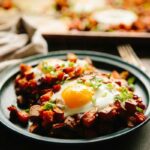 Plate of sweet potato and chorizo hash with eggs on top, garnished with scallions, with a sheet pan in the background.