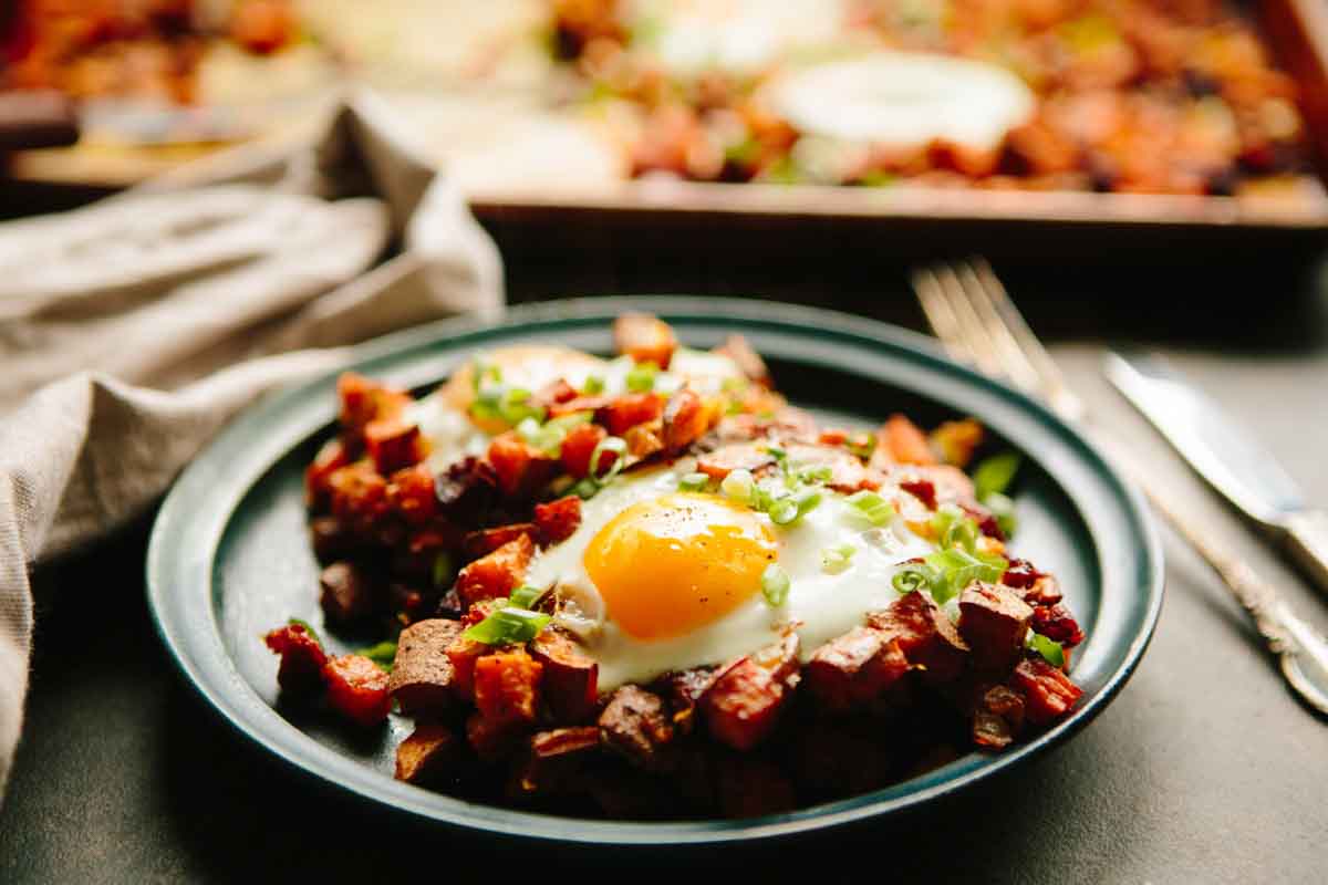 Plate of sweet potato and chorizo hash with eggs on top, garnished with scallions, with a sheet pan in the background.