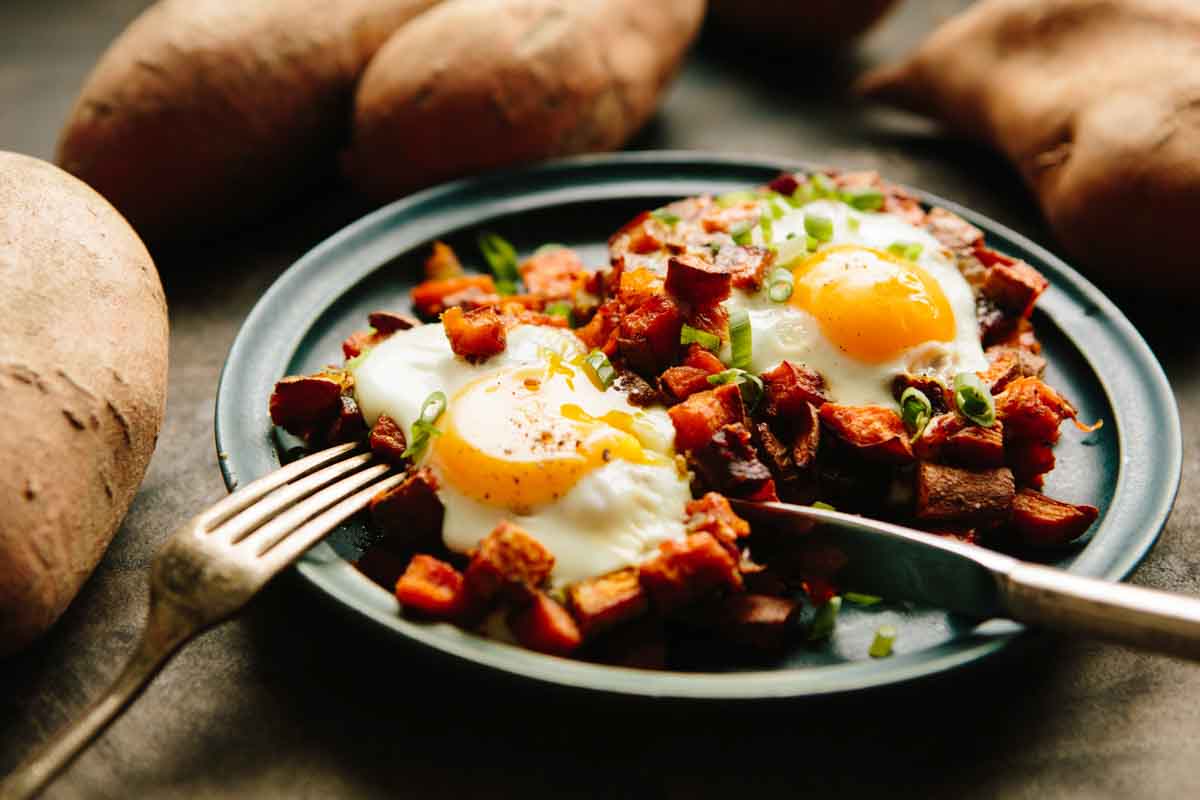 Two sunny-side-up eggs on sweet potato and chorizo hash with fresh scallions and whole sweet potatoes in the background.