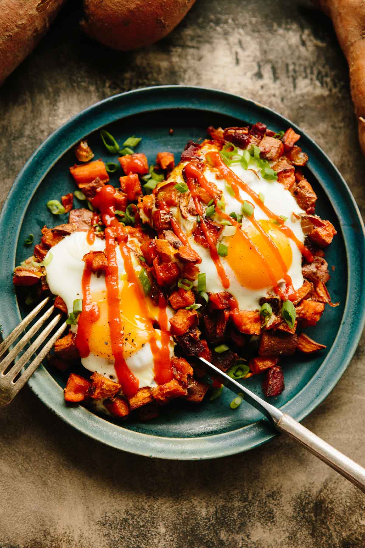 Sweet potato and chorizo hash topped with eggs and drizzled with sriracha on a blue plate.