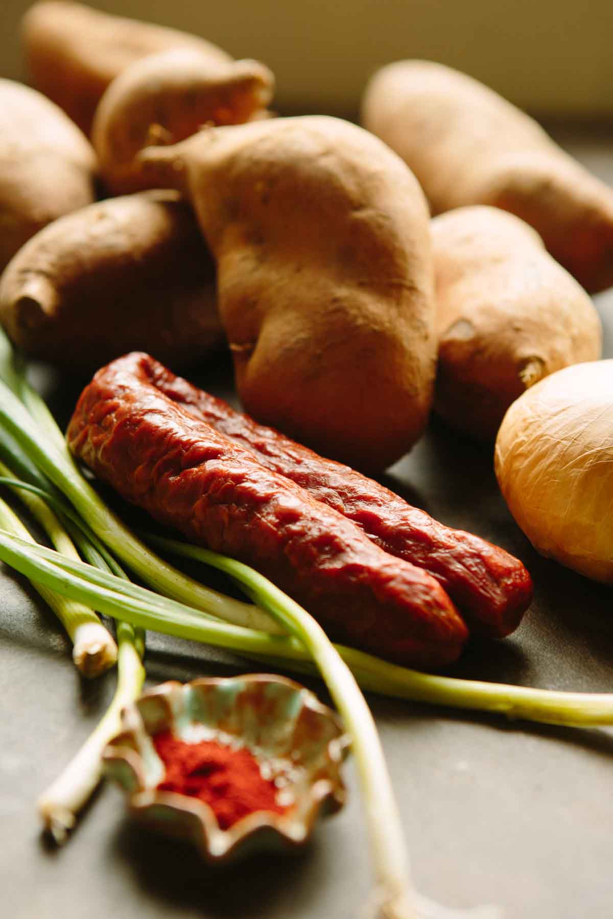 Close-up of raw ingredients including Spanish chorizo, sweet potatoes, scallions, onion, and a dish of paprika.