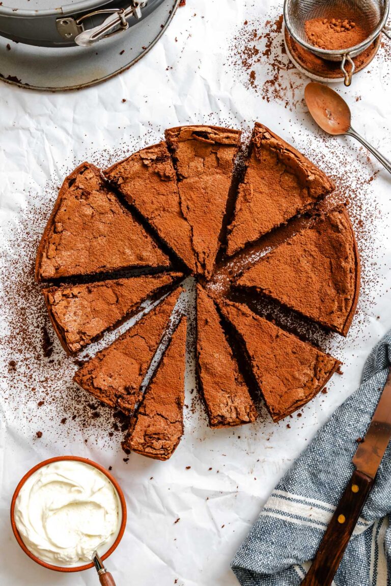 Overhead shot of sliced Torta Caprese dusted generously with cocoa powder, arranged on parchment with a bowl of whipped cream and a sieve nearby.
