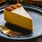 Slice of vegan pumpkin cheesecake on a dark blue plate with a gold fork, showing its creamy texture and nut crust.