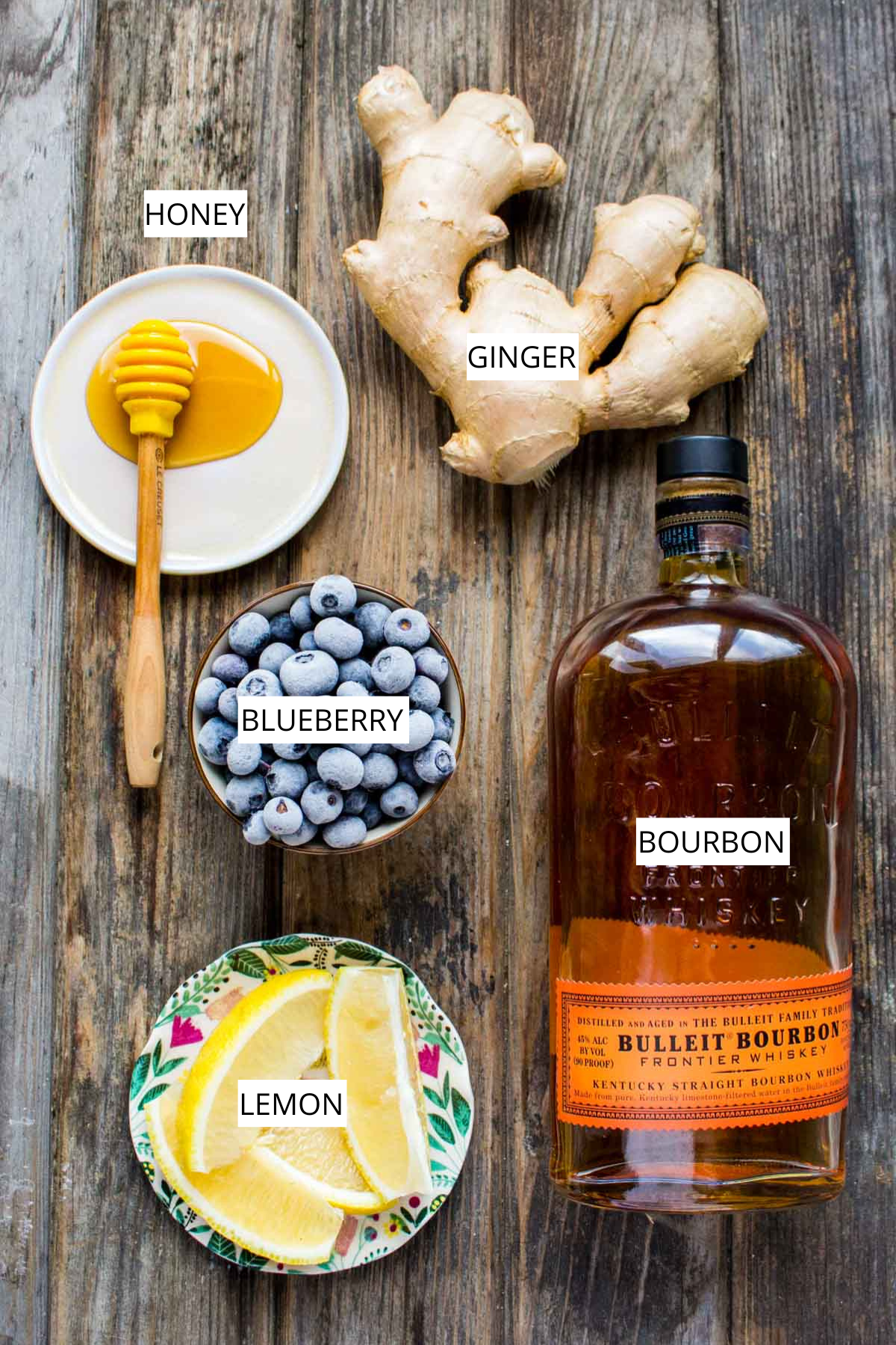 Overhead shot of labeled ingredients for a Blueberry Ginger Hot Toddy, including honey, ginger, blueberries, lemon, and a bottle of bourbon arranged on a wooden table.