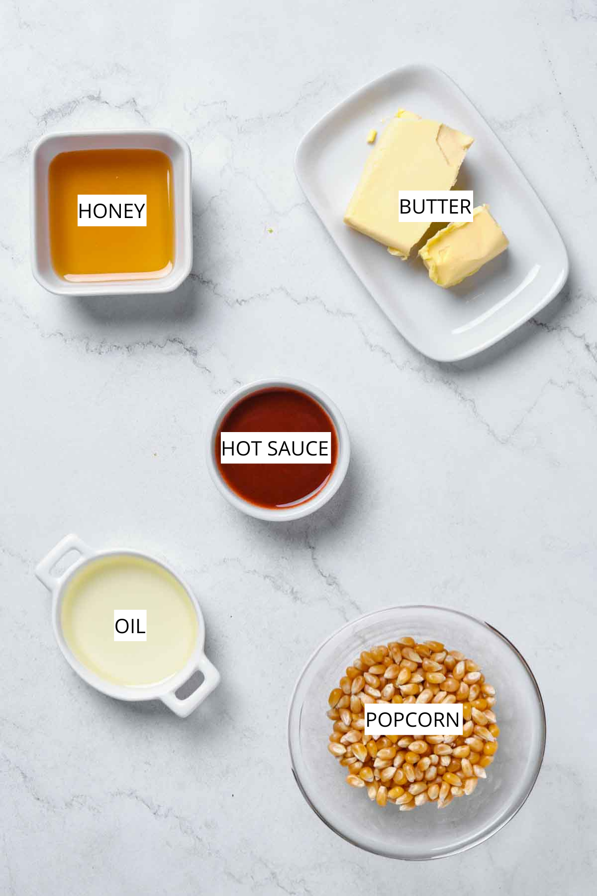 Overhead image of labeled ingredients: honey, butter, hot sauce, oil, and popcorn kernels on a white surface.