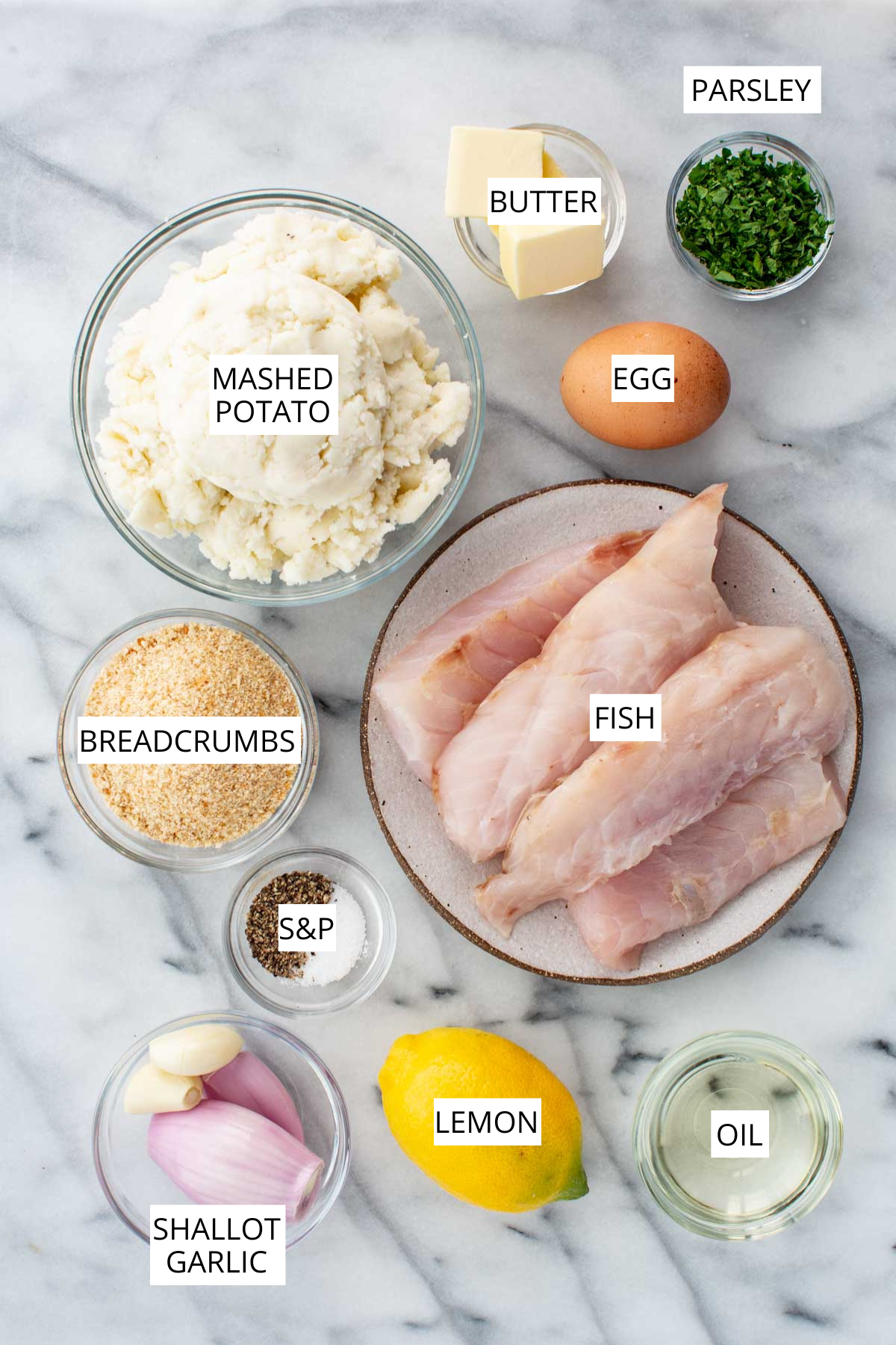 Overhead display of ingredients for mashed potato fish cakes including mashed potatoes, fish, breadcrumbs, butter, parsley, egg, shallot, garlic, lemon, oil and salt and pepper, each labeled.