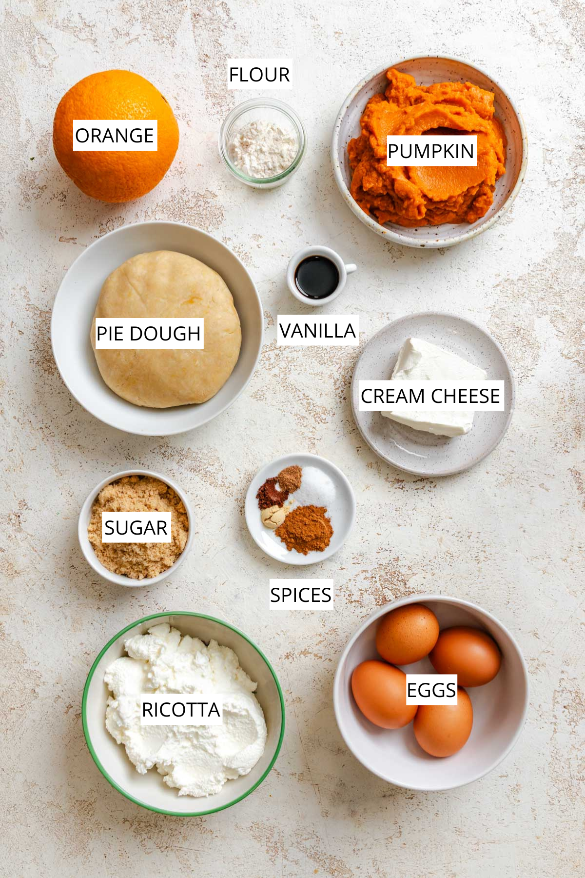 Overhead view of pumpkin ricotta pie ingredients arranged on a light surface, including ricotta, pumpkin purée, eggs, brown sugar, cream cheese, orange, vanilla, flour, pie dough, and warm spices.