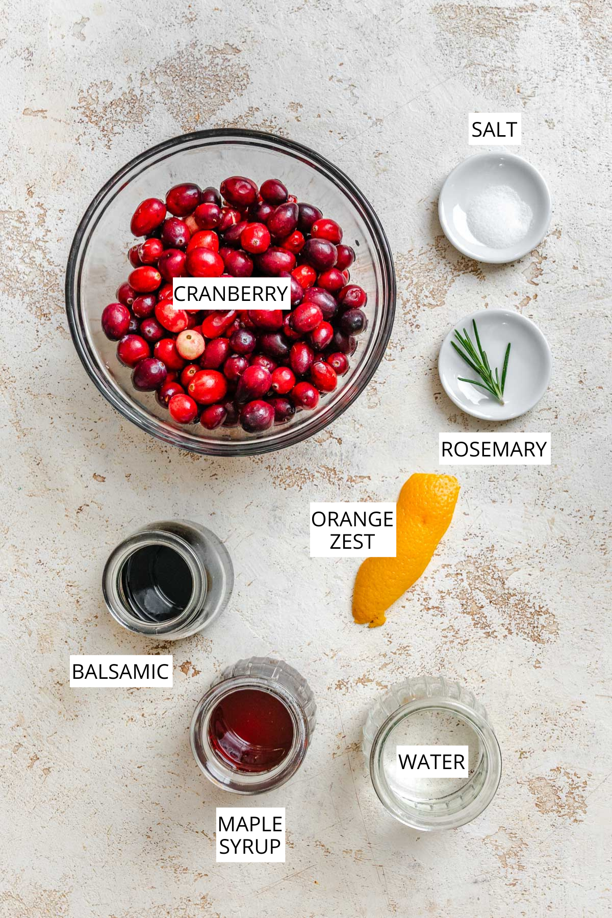 Overhead shot of ingredients for balsamic cranberry sauce - fresh cranberries, orange zest, rosemary, balsamic vinegar, maple syrup, water, and salt, arranged on a light surface and labeled.