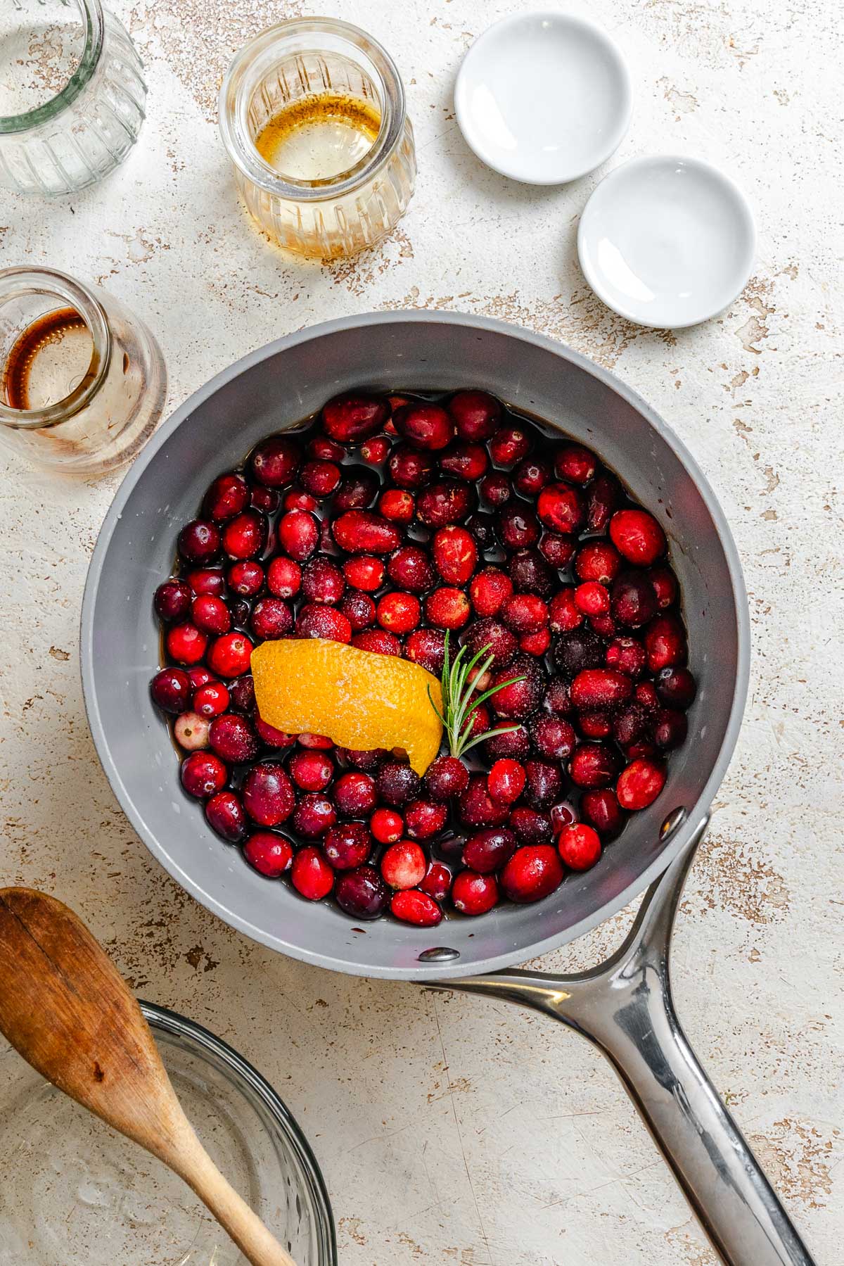 A saucepan filled with fresh cranberries, an orange peel, and a sprig of rosemary simmering with balsamic vinegar and maple syrup on a light textured surface.