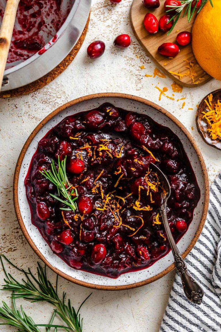 A serving bowl of glossy balsamic cranberry sauce topped with fresh orange zest and a sprig of rosemary, surrounded by cranberries and zest on the table.