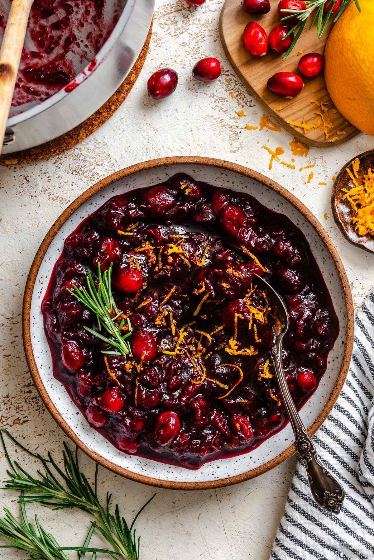 A serving bowl of glossy balsamic cranberry sauce topped with fresh orange zest and a sprig of rosemary, surrounded by cranberries and zest on the table.