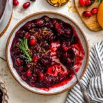 Overhead view of balsamic cranberry sauce in a rustic bowl with a spoon, garnished with orange zest and rosemary, next to a striped kitchen towel.
