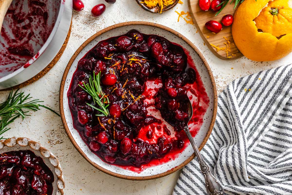 Overhead view of balsamic cranberry sauce in a rustic bowl with a spoon, garnished with orange zest and rosemary, next to a striped kitchen towel.