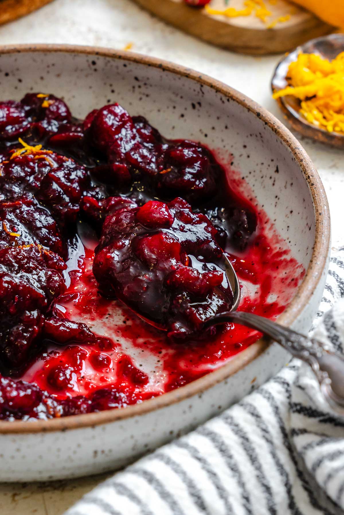 Side view of a spoon scooping the glossy cranberry balsamic sauce from a speckled ceramic bowl, with orange zest and cranberries in the background.