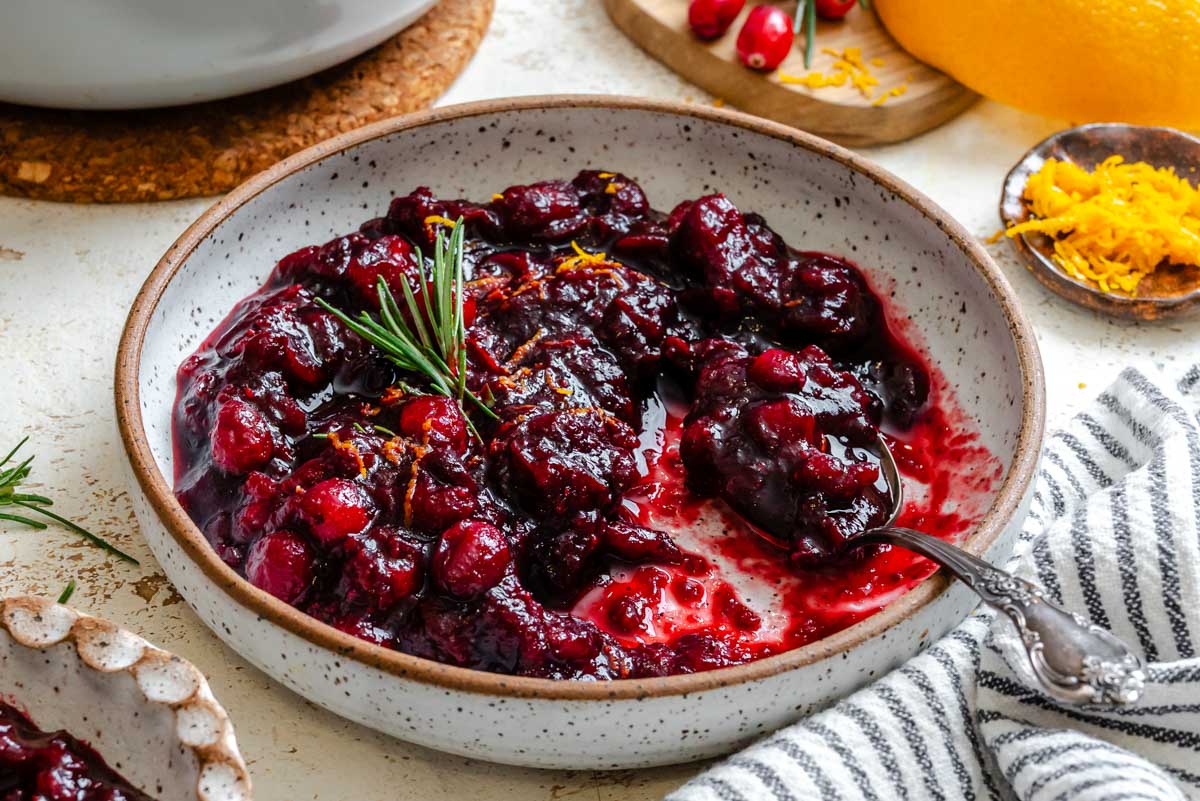 Bowl of rich, dark red cranberry sauce garnished with orange zest and rosemary, surrounded by ingredients like oranges and cranberries on a white table.