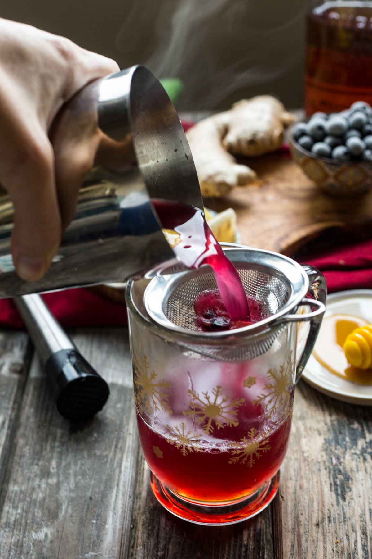 Pouring steaming blueberry and ginger mixture through a fine mesh strainer into a glass mug with gold snowflake designs, surrounded by honey, lemon, and ginger.