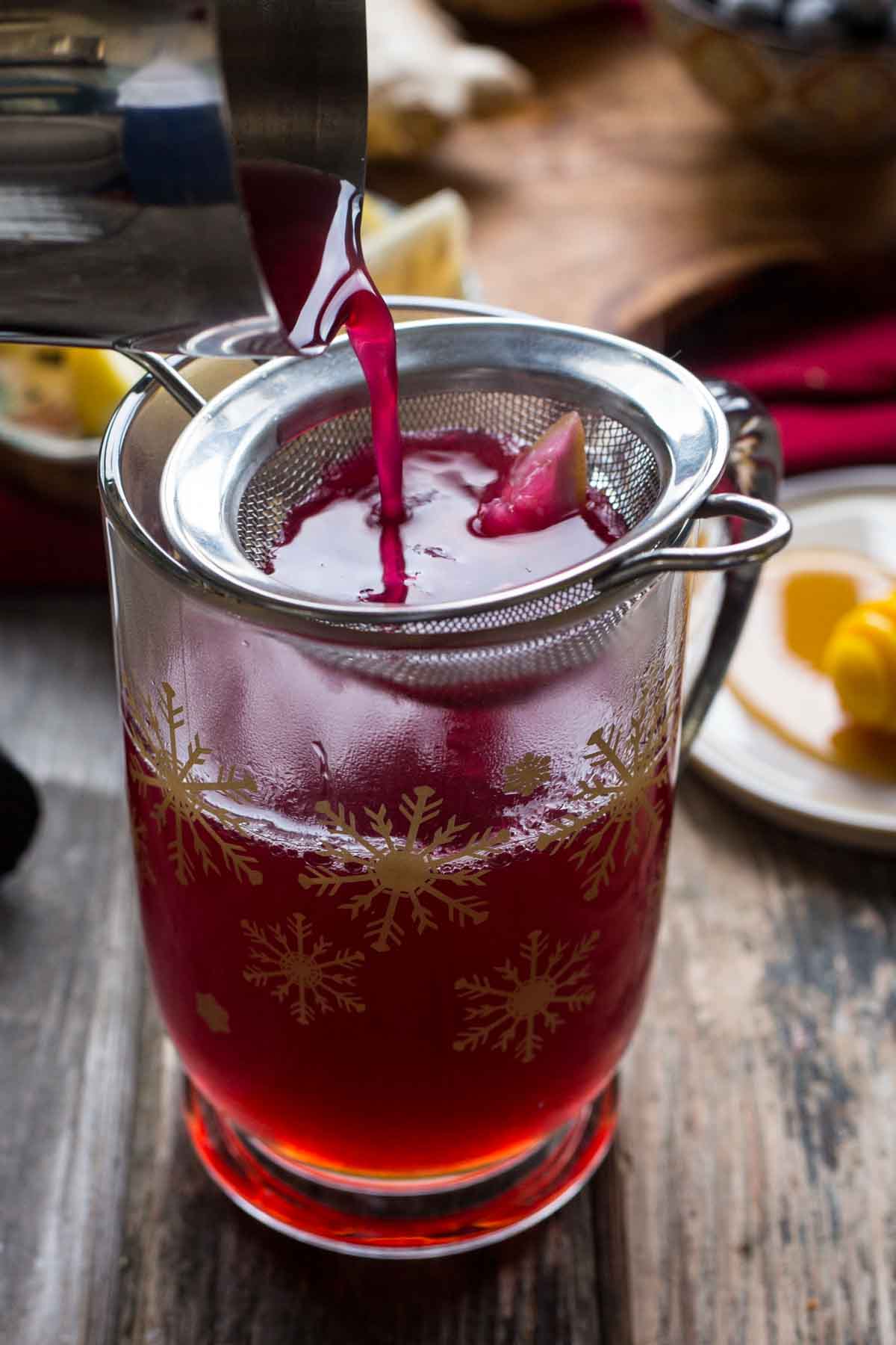 Close-up of vivid magenta blueberry ginger liquid being strained into a snowflake-patterned glass mug on a rustic wooden surface.