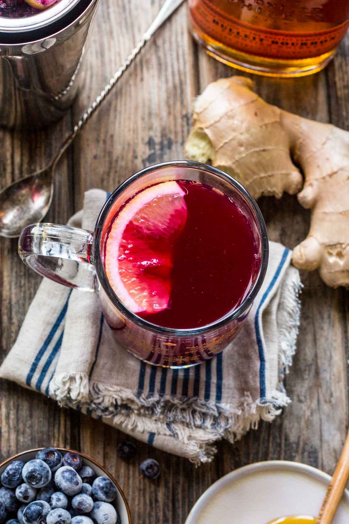 Overhead view of a Blueberry Ginger Hot Toddy with a lemon slice floating on top, surrounded by fresh ginger, blueberries, and honey on a wooden table.