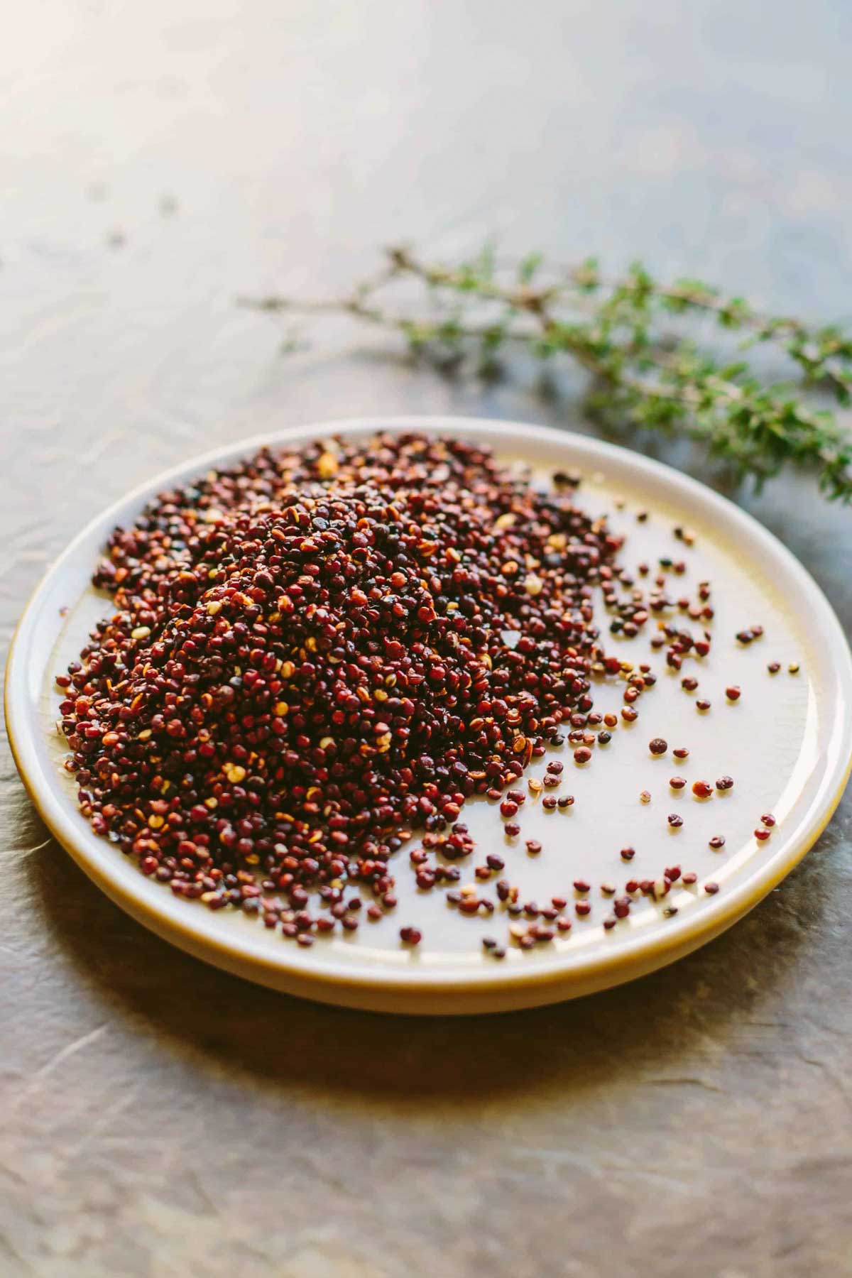 A small plate piled with toasted red quinoa, with loose grains scattered around and thyme sprigs in the background.