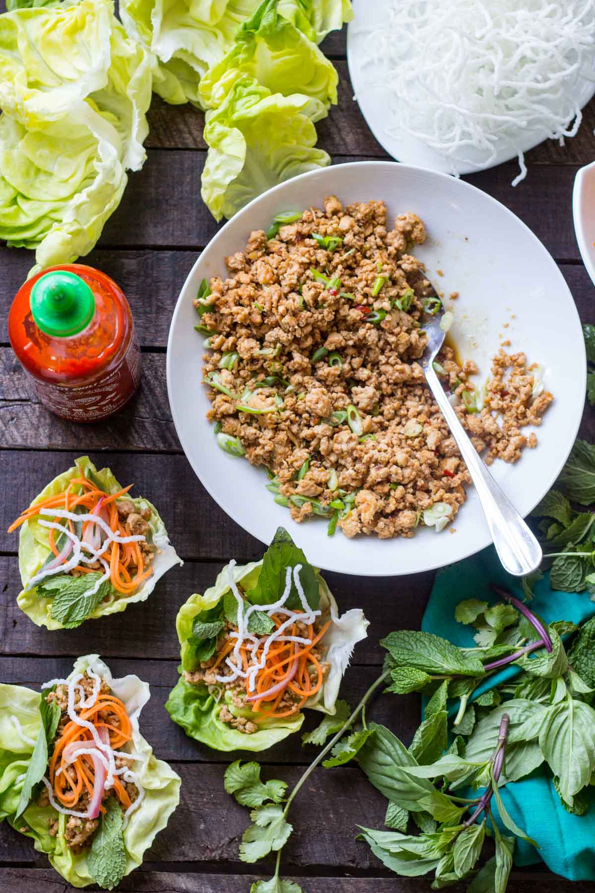 A table set with lettuce leaves, a large bowl of cooked chicken filling, fresh herbs, pickled vegetables and crispy rice noodles ready for assembling wraps.