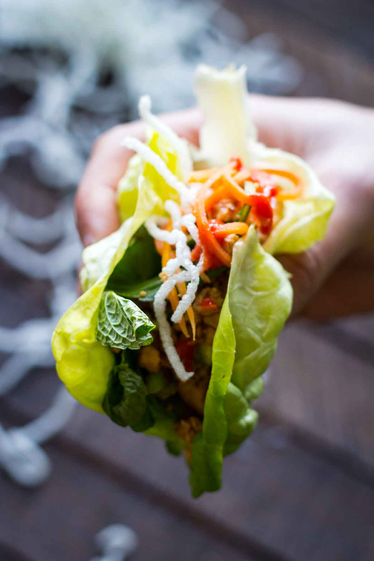 A hand holding a filled lettuce wrap with chicken, herbs, pickled carrots and puffed rice noodles.
