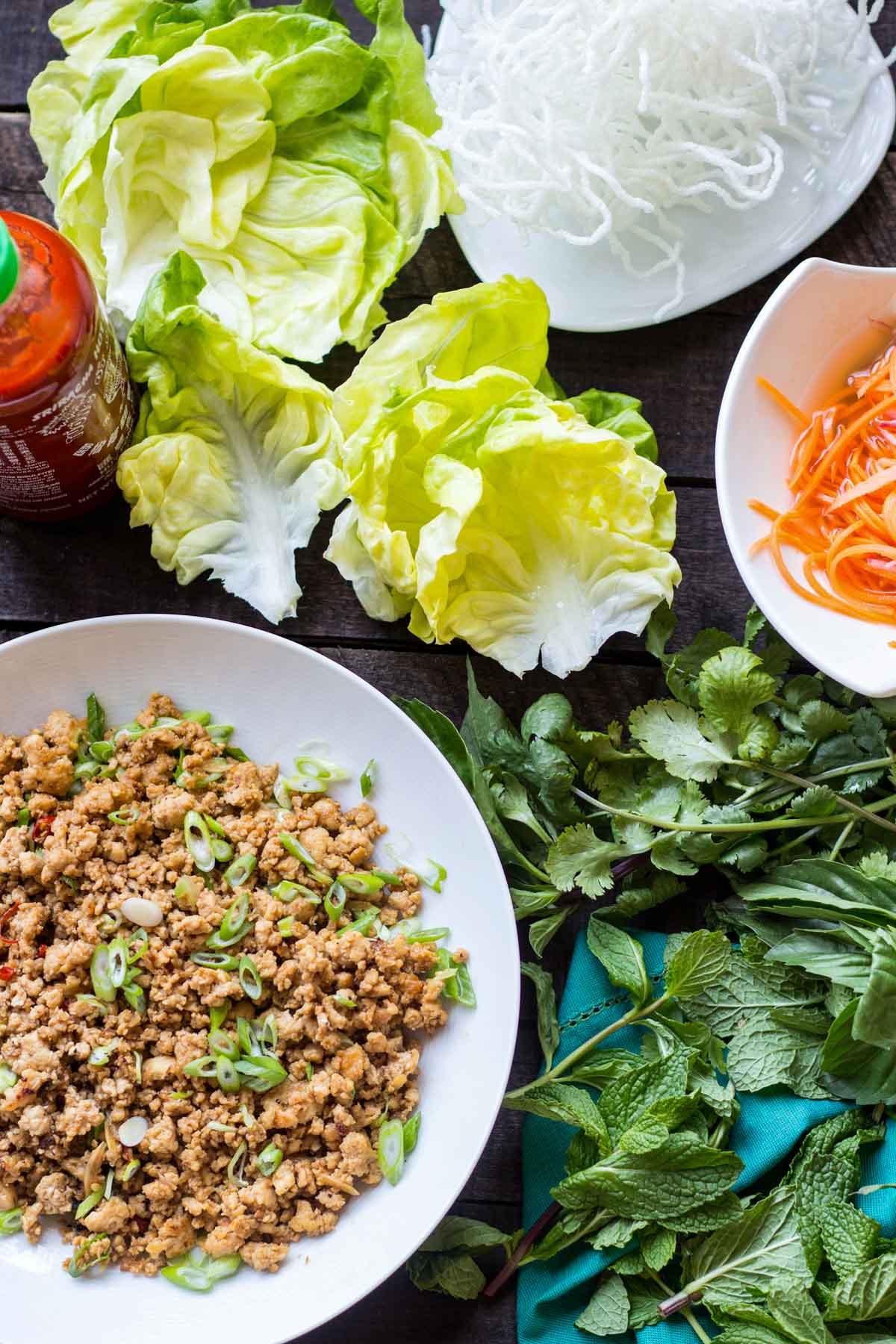 Lettuce leaves, a bowl of chicken filling, a plate of puffed rice noodles, fresh herbs and pickled vegetables arranged on a rustic wooden table.