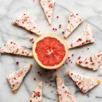 A halved ruby red grapefruit in the center with iced grapefruit shortbread triangles arranged in a circular pattern around it, sprinkled with pink peppercorns.
