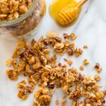 Crunchy homemade granola scattered on a marble surface next to a glass jar and a honey dipper resting in a small pool of honey.