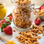 A clear latch-top jar overflowing with golden homemade granola surrounded by fresh strawberries and scattered clusters on a marble countertop.