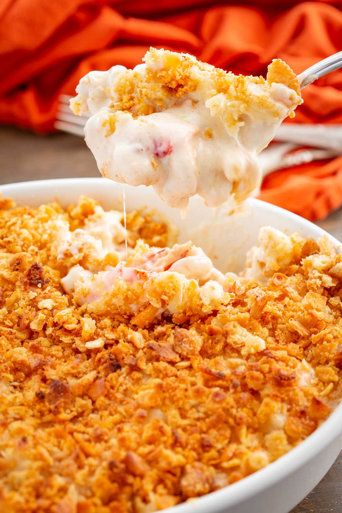 A close up of a spoon lifting a creamy scoop of lobster mac and cheese with a crunchy Ritz cracker topping from the casserole dish.