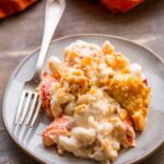 A plate of lobster mac n cheese showing large pieces of lobster, creamy pasta, and crisp cracker topping with a fork on the side.