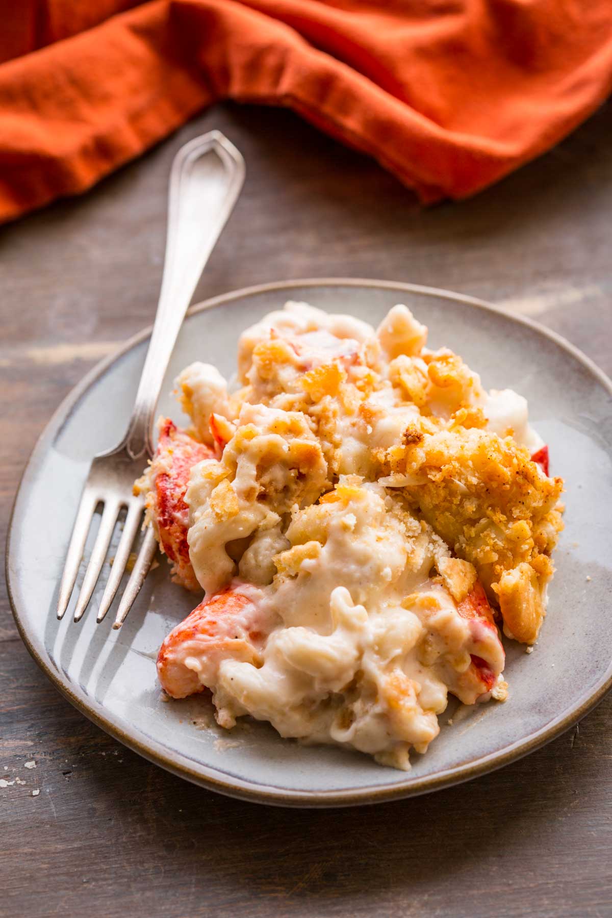 A plate of lobster mac and cheese showing large pieces of lobster, creamy pasta, and crisp cracker topping with a fork on the side.
