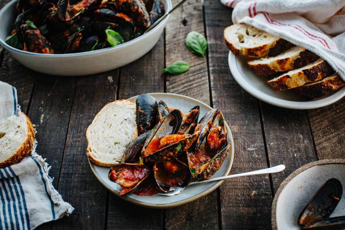 A small plate of mussels marinara served with crusty bread, surrounded by extra mussels and bread on a rustic wooden table.