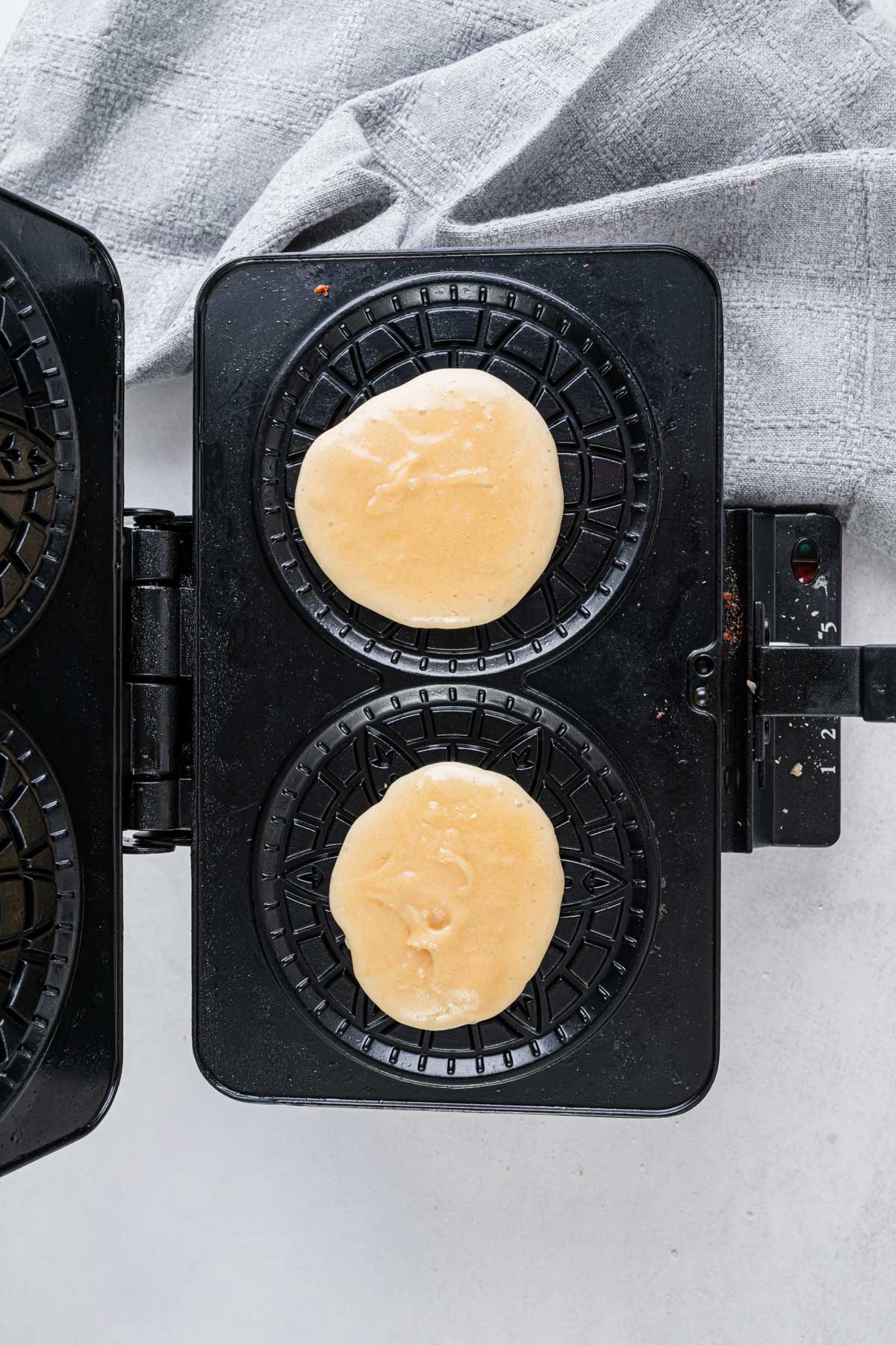 Two small portions of pizzelle batter placed on the patterned plates of a heated pizzelle iron.