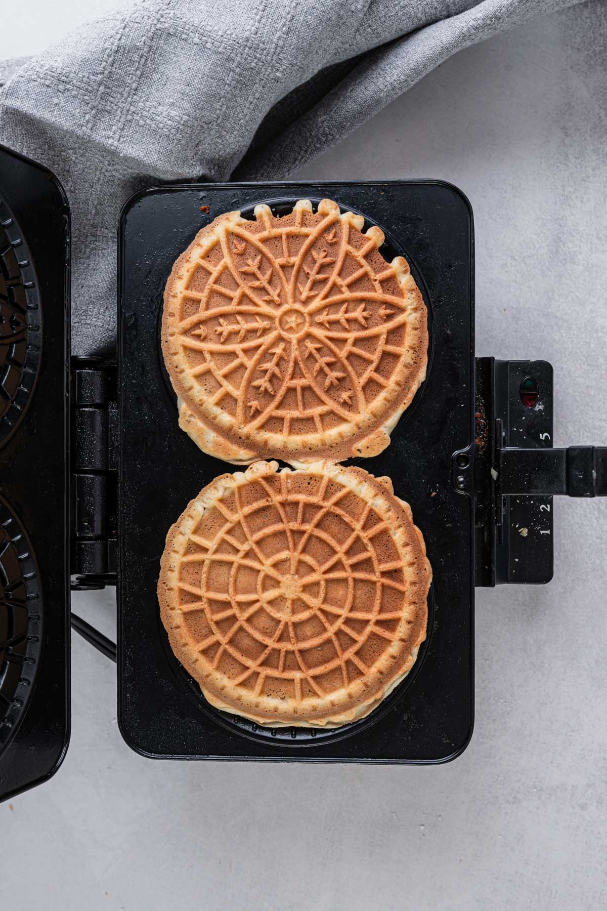 Two hot pizzelles with golden patterns still sitting on the open pizzelle iron after cooking.