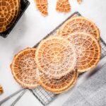 Golden pizzelles cooling on a wire rack, lightly dusted with powdered sugar, with broken cookie pieces around them.