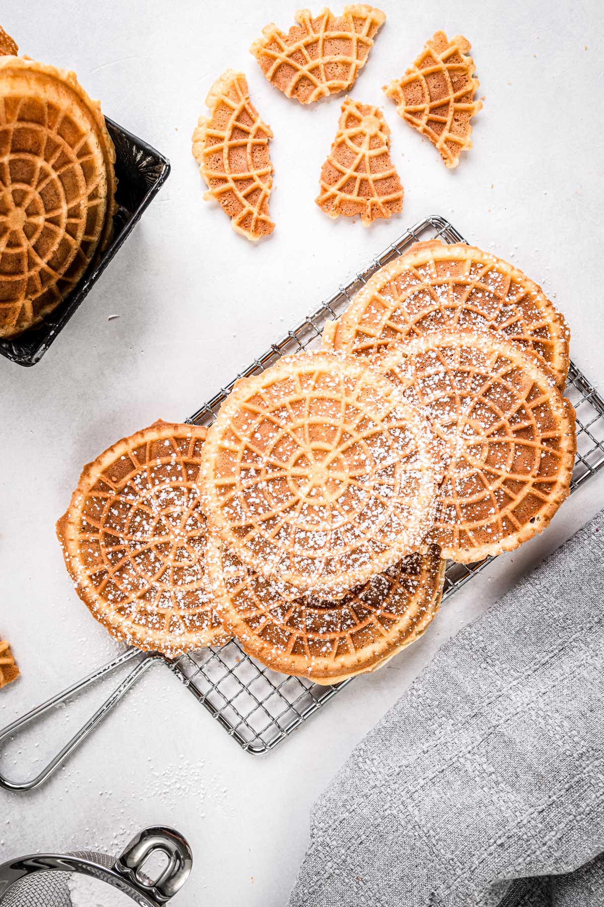 Golden pizzelles cooling on a wire rack, lightly dusted with powdered sugar, with broken cookie pieces around them.