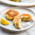 Two mashed potato fish cakes on a plate with one broken open to show the flaky interior, with a lemon wedge and fork on the side.