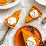 Slices of pumpkin ricotta pie served with whipped cream on an orange plate and parchment background, with forks nearby.