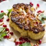 Pan-seared cauliflower steak served on a white plate over creamy tahini sauce, topped with pomegranate seeds, pine nuts, and fresh cilantro.
