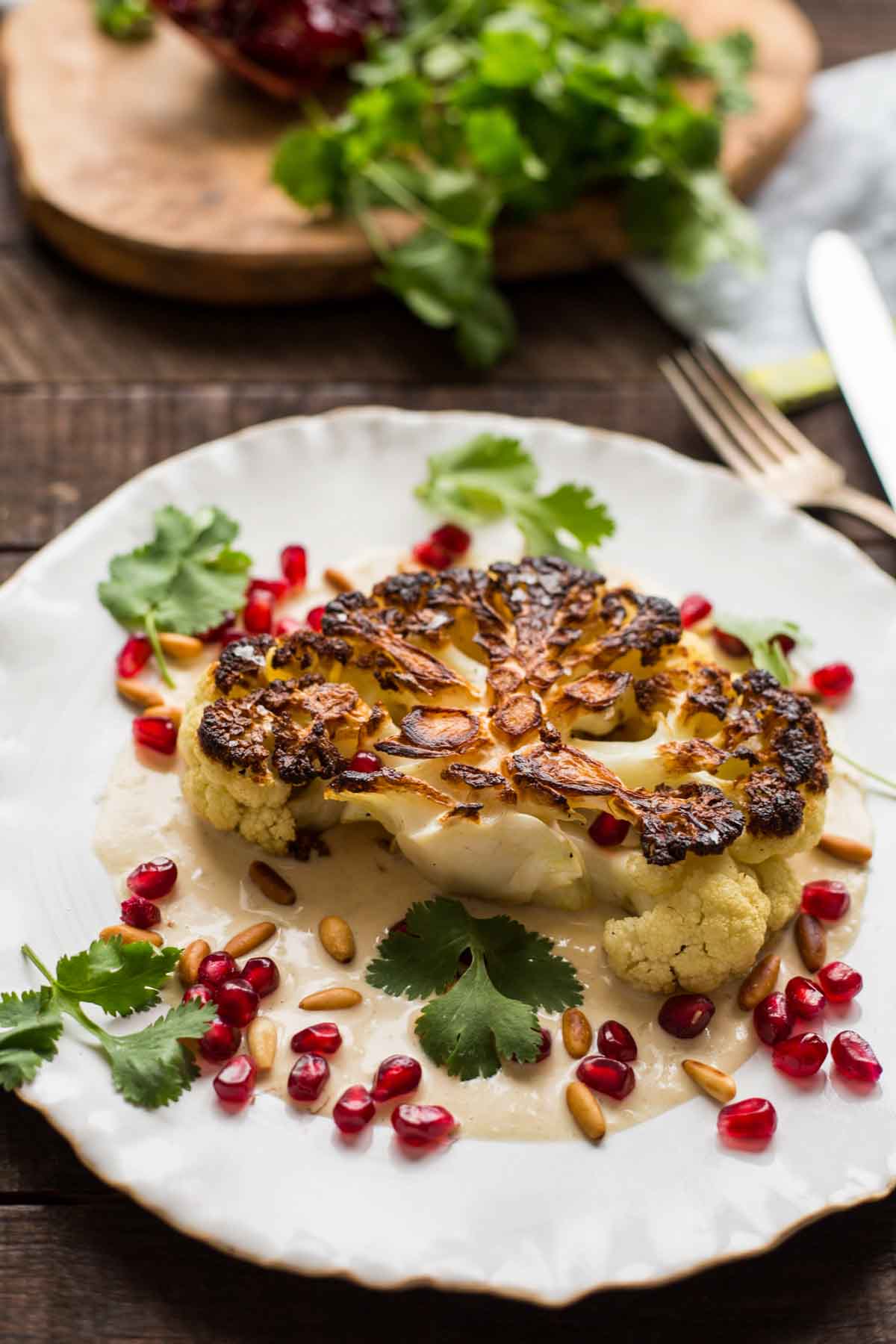 Overhead view of pan-seared cauliflower steak plated with tahini sauce, pine nuts, and bright red pomegranate seeds, styled on a rustic wooden table.