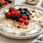 Bowl of thick strained Greek yogurt topped with fresh berries, honey, and granola on a speckled plate.
