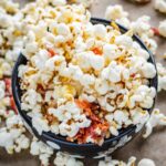 An overhead view of popcorn tossed with crisp prosciutto pieces, piled high in a black bowl with scattered kernels around it.