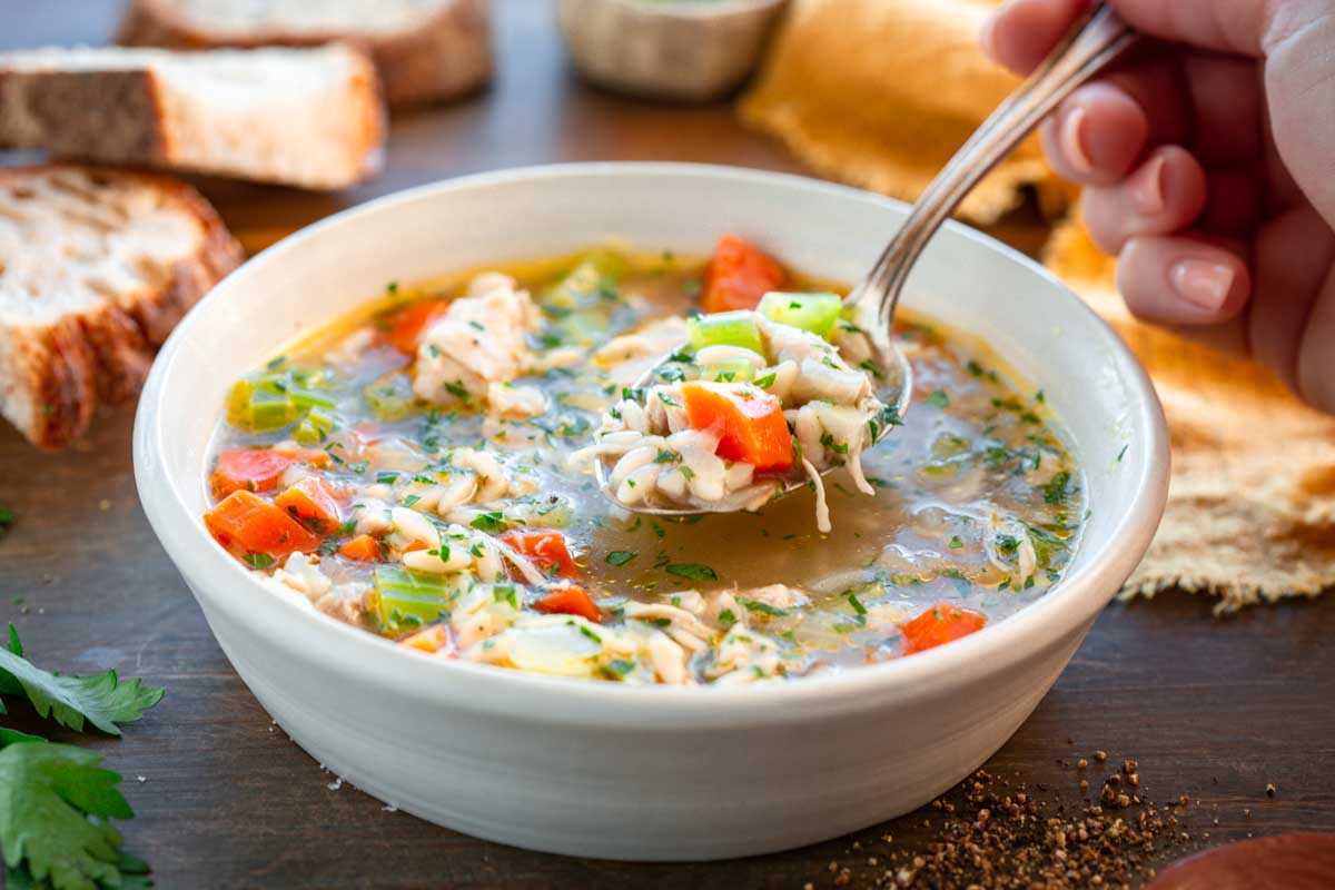 A hand lifts a spoonful of turkey, orzo and vegetables from a bowl of turkey orzo soup with bread in the background.