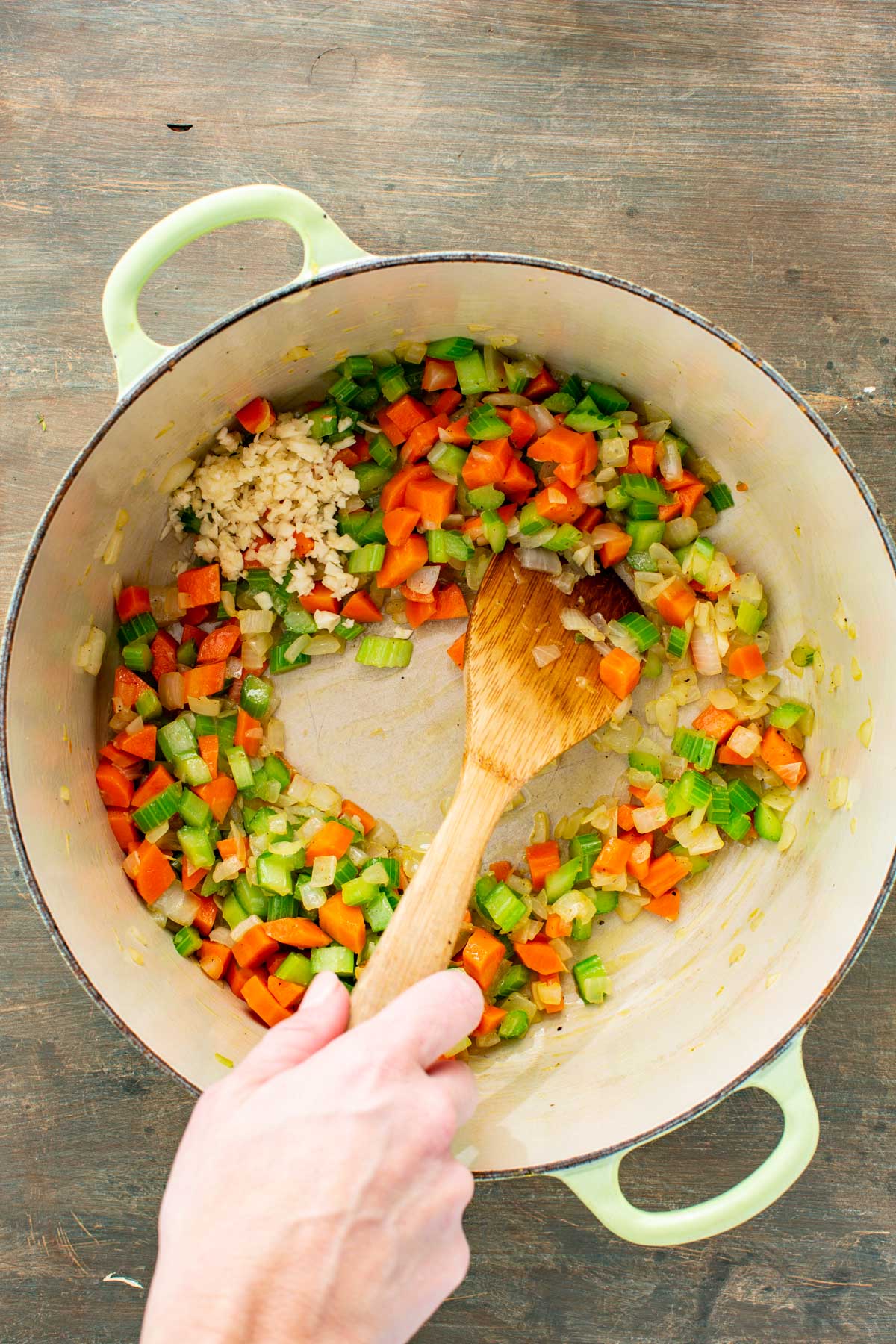 Carrots, celery and onions softening in a Dutch oven while minced garlic is added to the side.