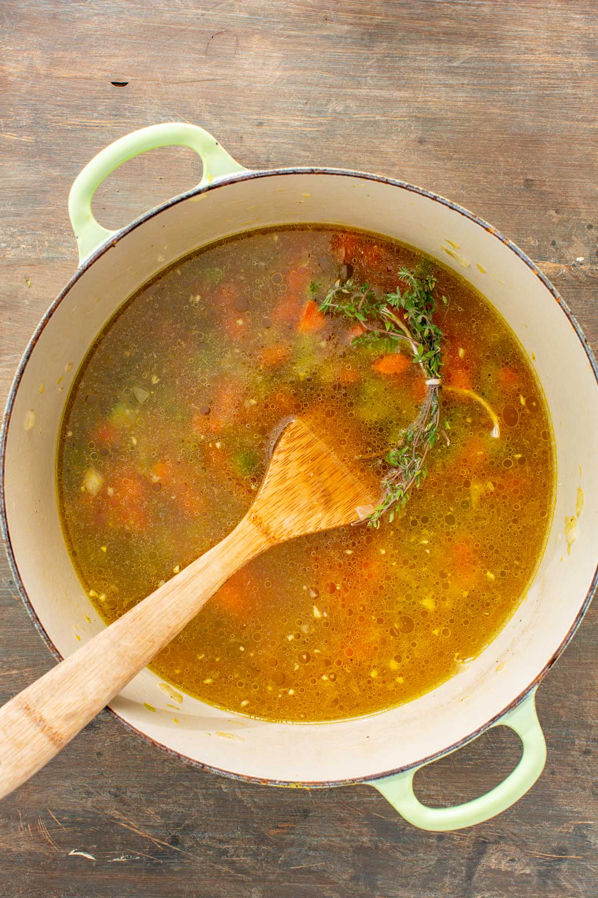 Broth and thyme simmering with sautéed vegetables in a Dutch oven, wooden spoon resting inside.